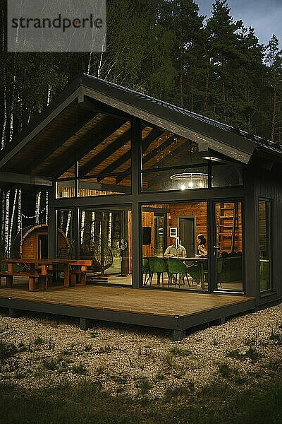 A view from outside at the couple sitting behind dining table in modern forest cabin in the evening