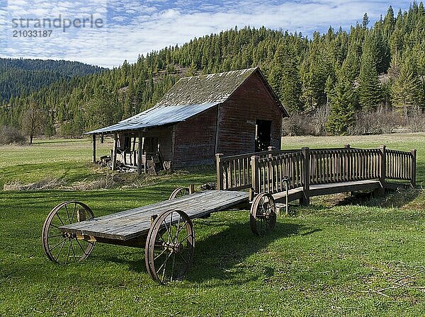 An old wagon sits near a small bridge and a small barn in north Idaho