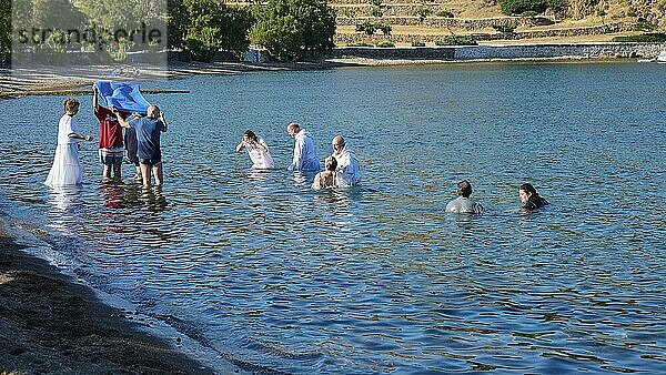 Men in white clothes and swimming costumes performing a ritual ceremony on the shore of a body of water in a hilly  sunny setting  baptism in the sea  Meli beach  Patmos  Dodecanese  Greek Islands  Greece  Europe