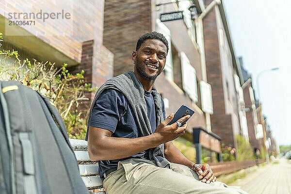 Happy african man using mobile sitting on a street bench next to a bag