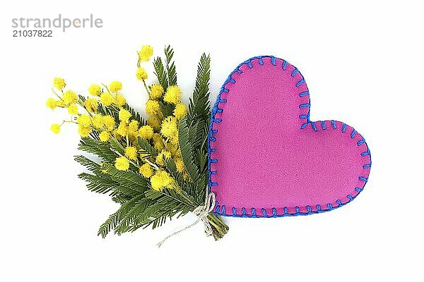 Heart-shaped pink pillow beside of yellow mimosa flowers against a white background  representation of a heartfelt sentiment expressed through the language of colors and shapes