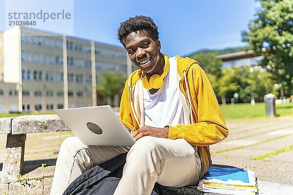 African american student using laptop sitting on the campus while smiling at camera