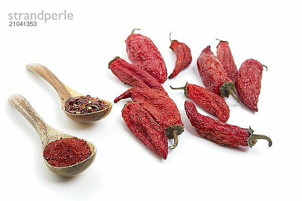 Red pepper powder and flakes on wooden spoons next to whole red peppers on a white background