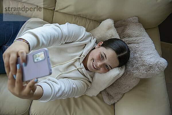 Overhead view of a young Caucasian woman lying on a sofa  capturing a selfie. Moment of joy and self-expression in a homely environment