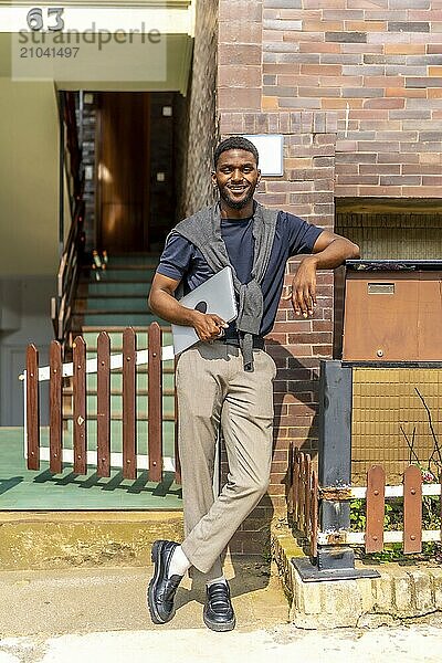 Vertical full length portrait of an african young businessman carrying a laptop outdoors next to a residential building