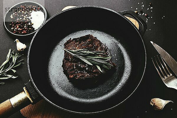 Cooked ribeye steak on cast-iron pan and board with pepper  rosemary  salt  garlic  knife  fork  butter on black background