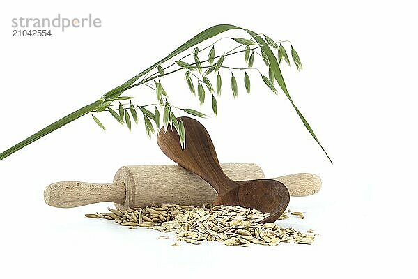 Oats groats rolled and flattened into flakes in wooden spoon and oats grains with hulls or husks near wooden rolling pin isolated on white background