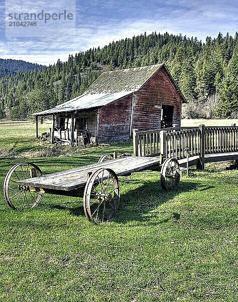 An old wagon sits near a small bridge and a small barn in north Idaho