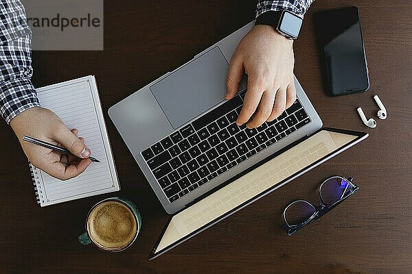 Man working at the laptop with a cup of coffee  phone  writing with pen at home.