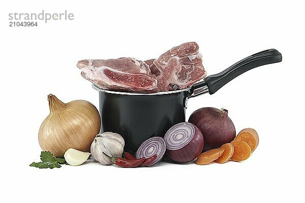 Black saucepan with pieces of raw pork inside. Around the saucepan are sliced vegetables which include onions  carrots  and garlic isolated on white background. Preparation of meat broth