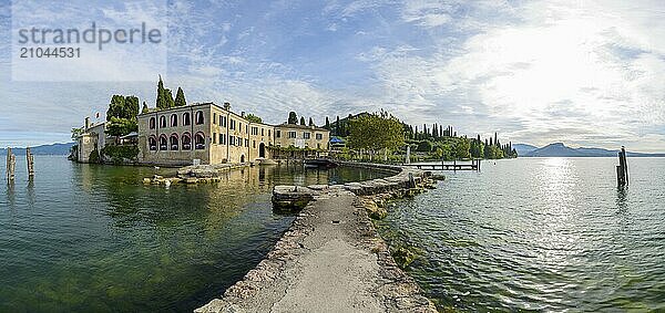 A serene view of a historic lakeside building by a calm lake with a pier extending into the water  surrounded by trees and mountains under a clear sky  Locanda San Vigilio  Punta San Vigilio  Garda  Lake garda  Lago di Garda  Veneto  Italy  Europe