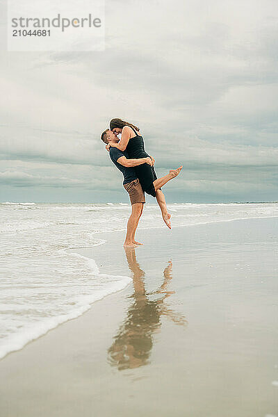 Man lifting woman on the beach as they kiss  with their reflection