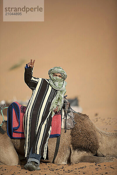 A Berber guide boy with a head scarf flashes a peace sign in front of a camel.