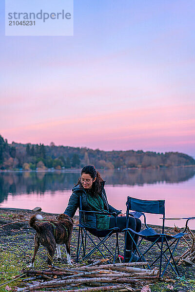 Woman playing with a dog with a wooden stick near a lake at sunset