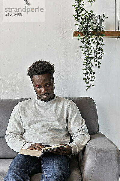 African American man reads book sitting on couch alone