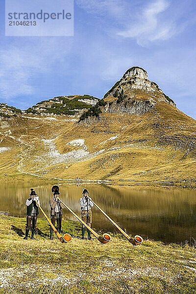 Drei Männer spielen Alphorn am Augstsee auf dem Berg Loser. Das österreichische Alphornbläser-Trio Klangholz. Im Hintergrund der Berg Atterkogel. Herbst  gutes Wetter  blauer Himmel. Spiegelung. Altaussee  Bad Aussee  Ausseer Land  Totes Gebirge  Steiermark  Oberösterreich  Österreich  Europa