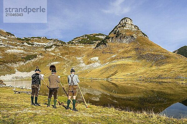 Drei Männer spielen Alphorn am Augstsee auf dem Berg Loser. Das österreichische Alphornbläser-Trio Klangholz. Im Hintergrund der Berg Atterkogel. Herbst  gutes Wetter  blauer Himmel. Spiegelung. Altaussee  Bad Aussee  Ausseer Land  Totes Gebirge  Steiermark  Oberösterreich  Österreich  Europa