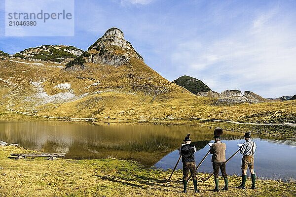 Drei Männer spielen Alphorn am Augstsee auf dem Berg Loser. Das österreichische Alphornbläser-Trio Klangholz. Im Hintergrund der Berg Atterkogel. Herbst  gutes Wetter  blauer Himmel. Spiegelung. Altaussee  Bad Aussee  Ausseer Land  Totes Gebirge  Steiermark  Oberösterreich  Österreich  Europa