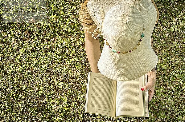 Hübsche Frau  die auf dem Rasen liegend ein Buch liest  von oben gesehen mit Sommerhut