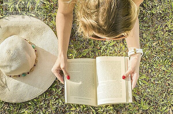 Hübsche Frau  die auf dem Rasen liegend ein Buch liest  von oben gesehen mit Sommerhut