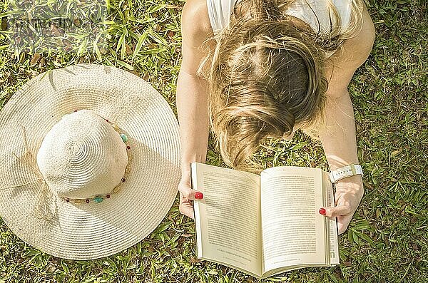 Hübsche Frau  die auf dem Rasen liegend ein Buch liest  von oben gesehen mit Sommerhut