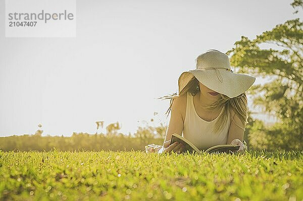 Hübsche Frau  die auf dem Rasen liegend ein Buch liest  von oben gesehen mit Sommerhut