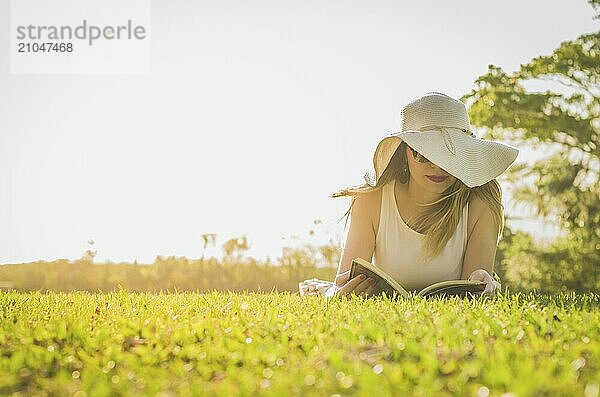 Hübsche Frau  die auf dem Rasen liegend ein Buch liest  von oben gesehen mit Sommerhut