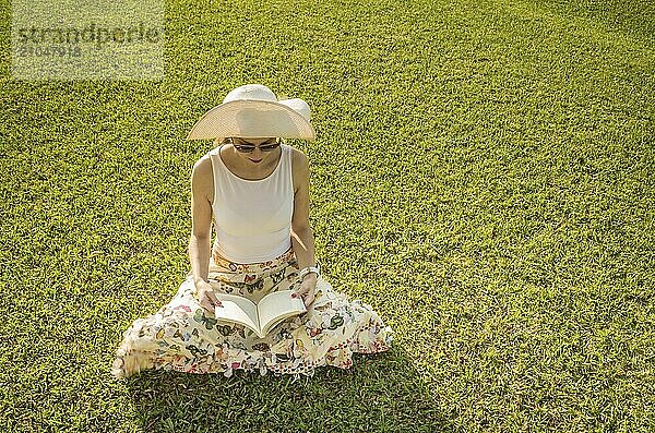 Hübsche Frau liest auf dem Rasen sitzend ein Buch  von oben gesehen mit Sommerhut