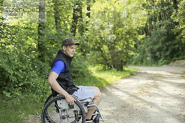 Glücklicher junger behinderter Mann in einem Rollstuhl sitzend in der Natur  der auf einem Wanderweg an einem schönen sonnigen Tag die Räder dreht