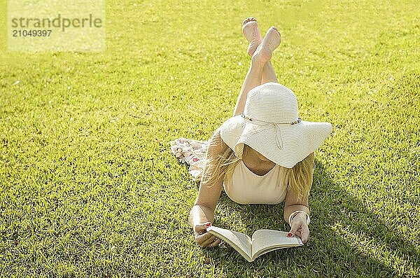 Hübsche Frau  die auf dem Rasen liegend ein Buch liest  von oben gesehen mit Sommerhut