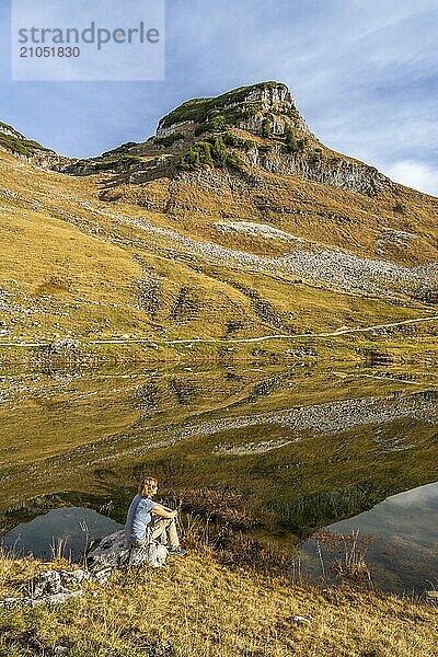 Augstsee und der Berg Atterkogel auf dem Loser. Eine Wanderin sitzt auf einem Felsen. Herbst  gutes Wetter  blauer Himmel. Spiegelung. Altaussee  Bad Aussee  Ausseer Land  Totes Gebirge  Steiermark  Oberösterreich  Österreich  Europa