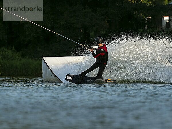 Junger Mann mit Wakeboard im See vor Schanze  Wassersport  Wasserski im Wakepark