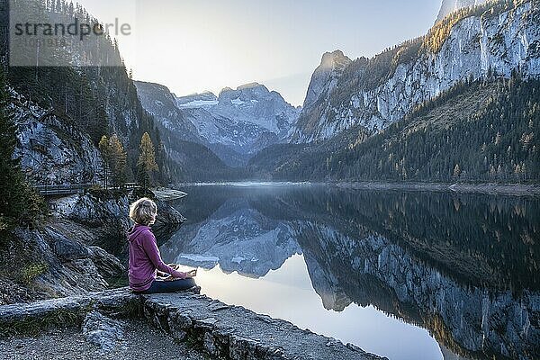 Eine Frau macht Yoga am See. Lotussitz (Padmasana) . Vorderer Gosausee im Herbst mit Blick auf das Gebirge des Dachstein. Rechts der Gosaukamm. Blauer Himmel  gutes Wetter. Spiegelung. Morgenstimmung. Vorderer Gosausee  Gosau  Gosautal  Salzkammergut  Oberösterreich  Österreich  Europa