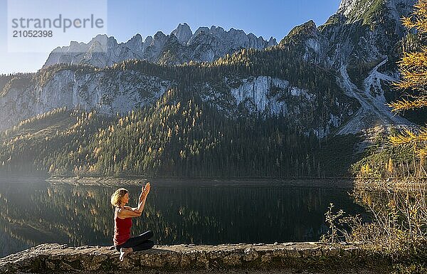 Eine Frau macht Yoga (Gomukhasana  Garudasana) am See. Vorderer Gosausee im Herbst mit Blick auf das Gebirge des Dachstein. Rechts der Gosaukamm. Blauer Himmel  gutes Wetter. Spiegelung. Morgenstimmung. Vorderer Gosausee  Gosau  Gosautal  Salzkammergut  Oberösterreich  Österreich  Europa