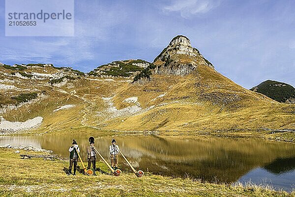 Drei Männer spielen Alphorn am Augstsee auf dem Berg Loser. Das österreichische Alphornbläser-Trio Klangholz. Im Hintergrund der Berg Atterkogel. Herbst  gutes Wetter  blauer Himmel. Spiegelung. Altaussee  Bad Aussee  Ausseer Land  Totes Gebirge  Steiermark  Oberösterreich  Österreich  Europa