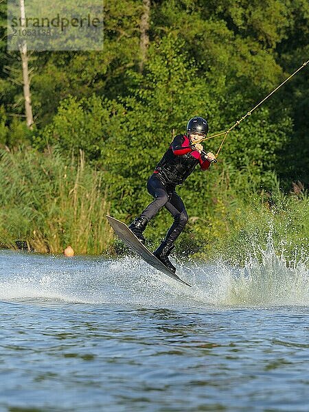 Junger Mann bei Sprung mit Wakeboard im See  Wassersport  Wasserski im Wakepark