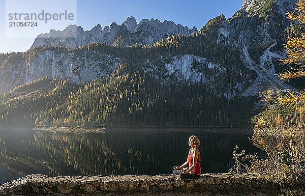 Eine Frau macht Yoga am See. Lotussitz (Padmasana) . Vorderer Gosausee im Herbst mit Blick auf das Gebirge des Dachstein. Rechts der Gosaukamm. Blauer Himmel  gutes Wetter. Spiegelung. Morgenstimmung. Vorderer Gosausee  Gosau  Gosautal  Salzkammergut  Oberösterreich  Österreich  Europa