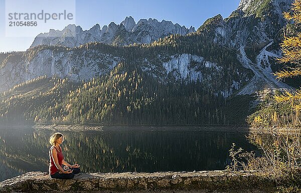 Eine Frau macht Yoga am See. Lotussitz (Padmasana) . Vorderer Gosausee im Herbst mit Blick auf das Gebirge des Dachstein. Rechts der Gosaukamm. Blauer Himmel  gutes Wetter. Spiegelung. Morgenstimmung. Vorderer Gosausee  Gosau  Gosautal  Salzkammergut  Oberösterreich  Österreich  Europa