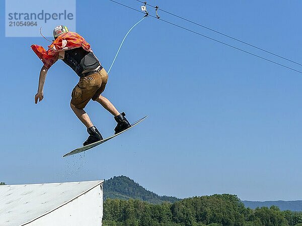 Junger lässiger Mann mit flatternden Hemd auf Wakeboard bei Sprung über Schanze in den See  Wasserski und Wakepark  Stráž pod Ralskem  Tschechin