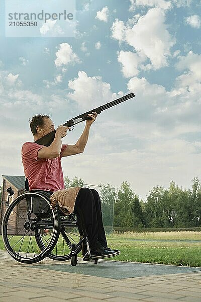 Portrait im Freien eines älteren Mannes im Rollstuhl  der ein Gewehr in den Himmel hält und auf ein Flugziel wartet