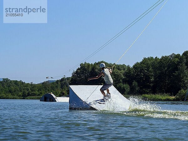 Junger Mann bei Sprung mit Wakeboard über Schanze in den See  Wassersport  Wasserski im Wakepark  Stráž pod Ralskem  Tschechin