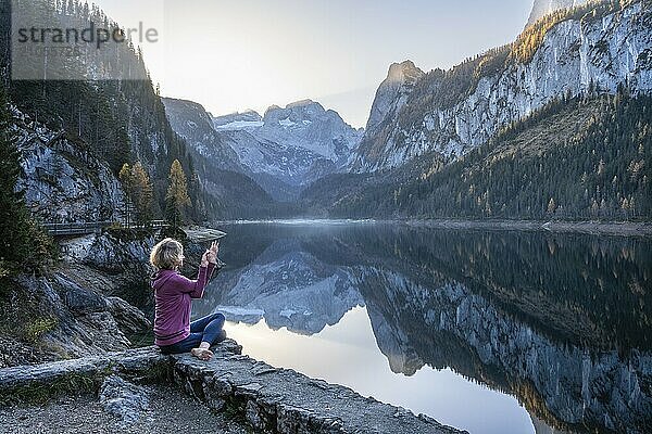 Eine Frau macht Yoga (Gomukhasana  Garudasana) am See. Vorderer Gosausee im Herbst mit Blick auf das Gebirge des Dachstein. Rechts der Gosaukamm. Blauer Himmel  gutes Wetter. Spiegelung. Morgenstimmung. Vorderer Gosausee  Gosau  Gosautal  Salzkammergut  Oberösterreich  Österreich  Europa