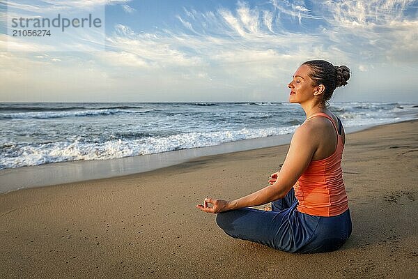 Junge sportliche  fitte Frau macht Yoga  meditiert und entspannt sich in Padmasana Lotus Pose) mit Kinn Mudra im Freien am tropischen Strand bei Sonnenuntergang