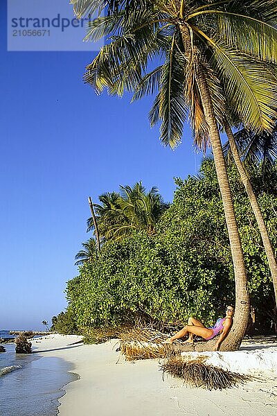 Junge Frau in bunter Badeanzug lehnt entspannt an schräg gewachsener Stamm von Kokospalme (Cocos nucifera) an Strand von Malediveninsel in Indischer Ozean  Malediven  Asien