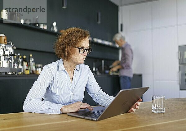 Symbolfoto zum Thema Arbeitsteilung bei Paaren im Haushalt. Eine Frau sitzt in einer Küche am Laptop während ein Mann im Hintergrund Geschirr spült. Berlin  13.08.2024