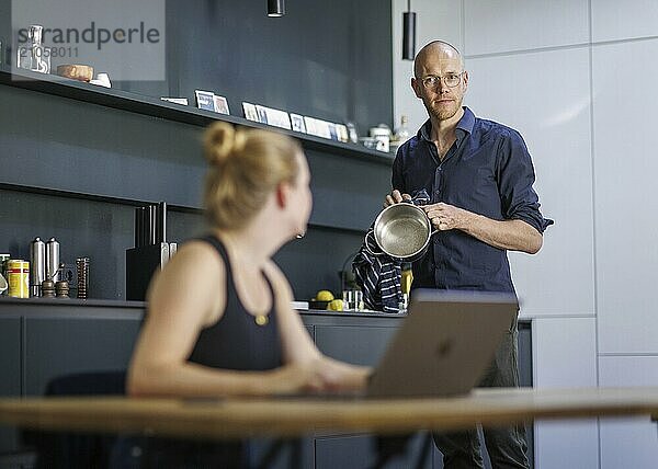 Symbolfoto zum Thema Arbeitsteilung bei Paaren im Haushalt. Eine Frau sitzt in einer Küche am Laptop während ein Mann im Hintergrund Geschirr spült. Berlin  13.08.2024