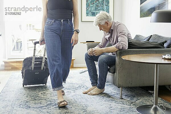 Symbolfoto zum Thema Trennung in der Partnerschaft. Eine Frau zieht einen Rollkoffer durch ein Wohnzimmer während ihr Mann deprimiert auf dem Sofa sitzt. Berlin  13.08.2024