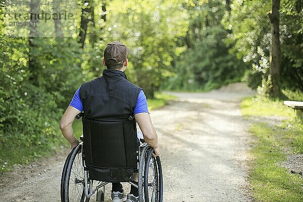 Glücklicher junger behinderter Mann in einem Rollstuhl sitzend in der Natur  der auf einem Wanderweg an einem schönen sonnigen Tag die Räder dreht