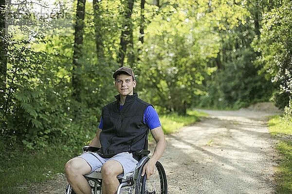 Glücklicher junger behinderter Mann in einem Rollstuhl sitzend in der Natur  der auf einem Wanderweg an einem schönen sonnigen Tag die Räder dreht