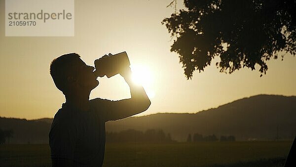 Silhouette eines fitten jungen Mannes  der Wasser trinkt  mit Sonne und Natur dahinter im Sonnenuntergang